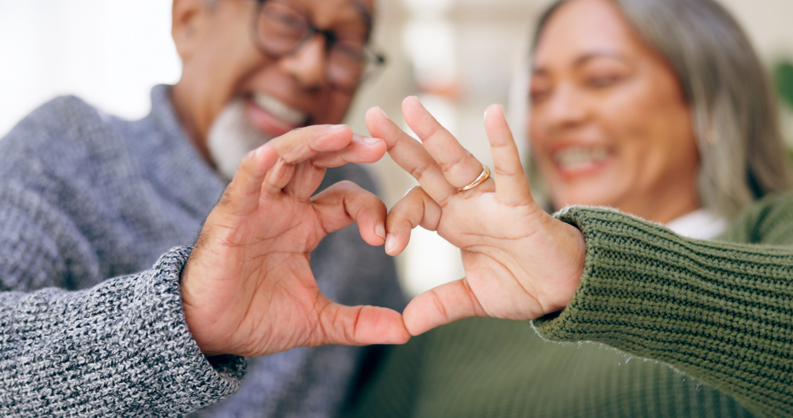 An older couple smiles together while forming a heart shape with their hands in the foreground.